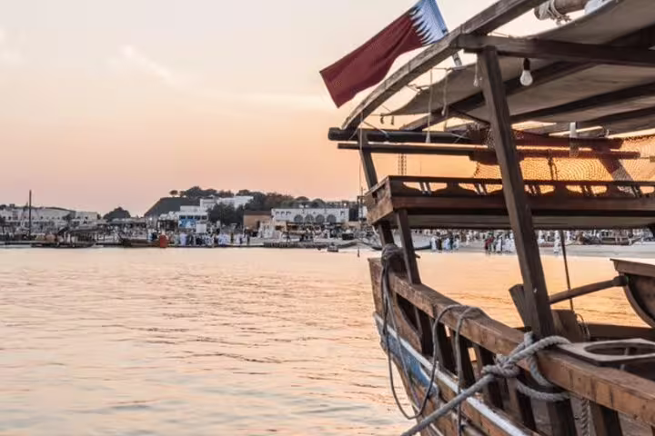 Traditional dhow boat with a Qatari flag at sunset in Doha, offering a scenic view of the waterfront and skyline.