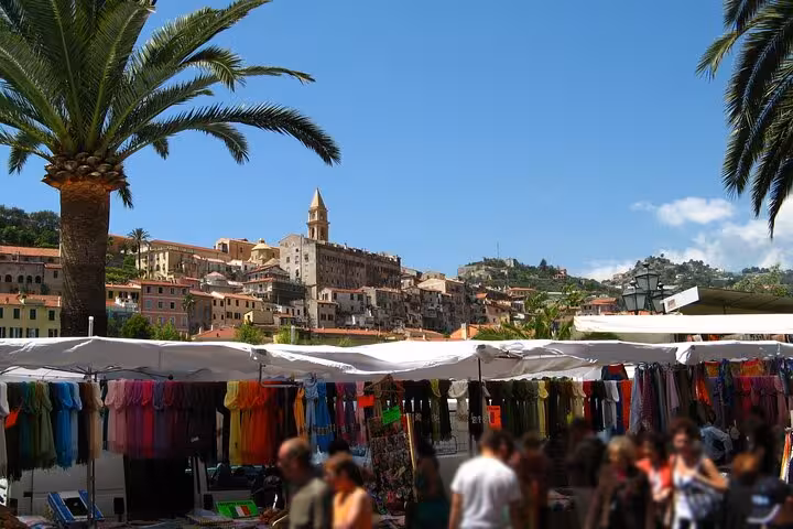 Colorful outdoor market stalls with clothing displays beneath palm trees and hilltop village of Dolceacqua, Italy