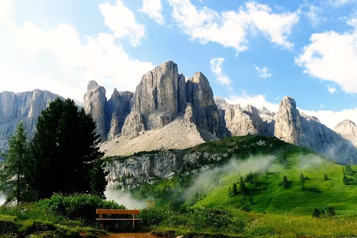 Majestic Dolomite peaks under a blue sky, overlooking a lush green landscape with a solitary bench and mist.