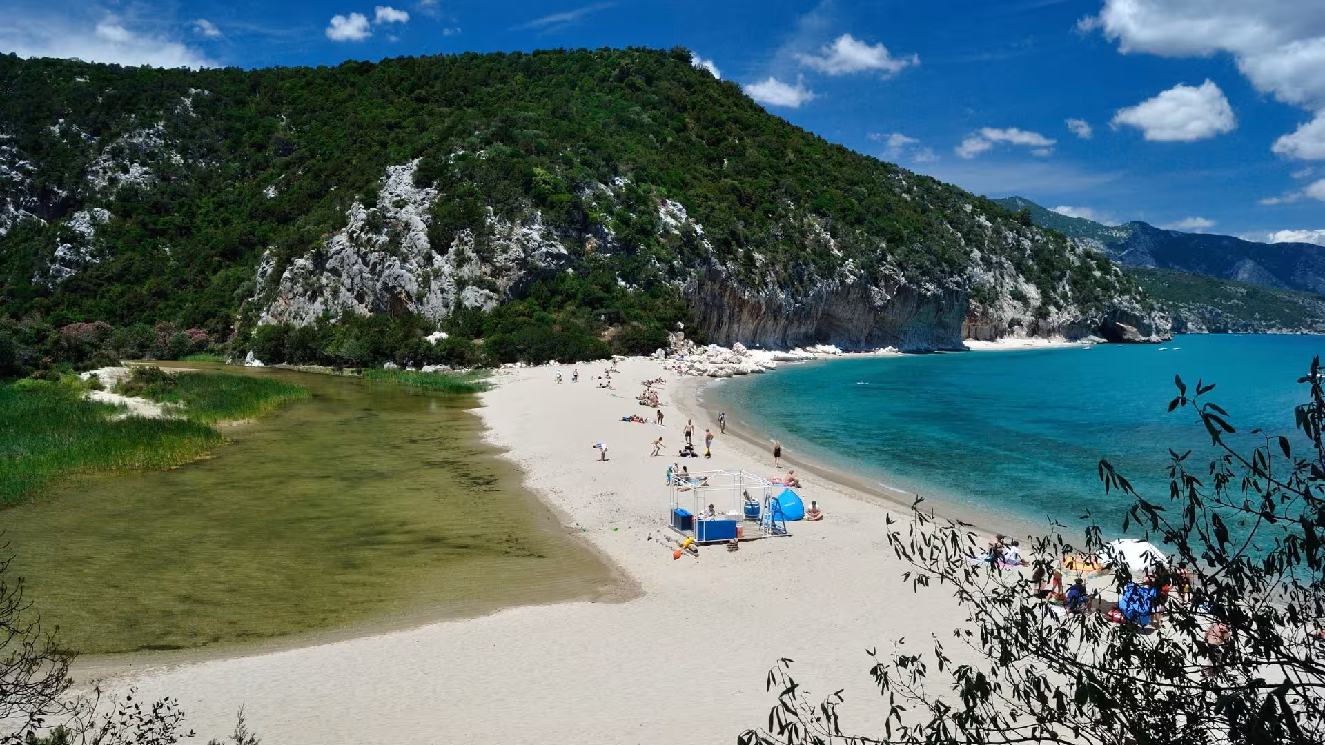 Tourists enjoying the sandy shore and clear waters at Cala Luna beach, surrounded by lush greenery and cliffs.
