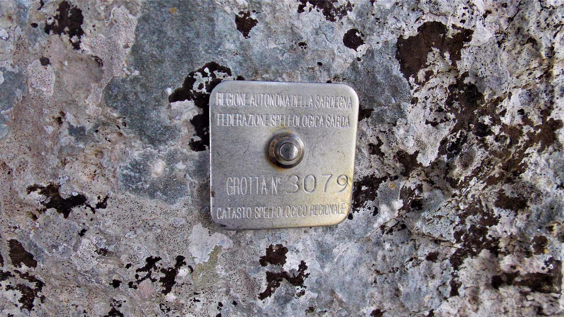 Metal plaque on a weathered rock in Dorgali marking a cave entrance for speleology enthusiasts.