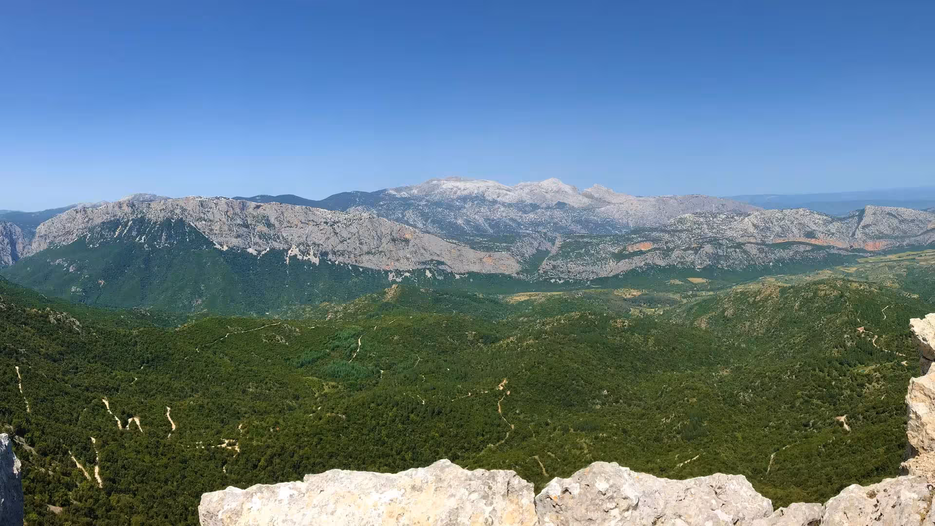 Expansive view of Sardinia's mountainous landscape with verdant valleys and rocky peaks under a clear blue sky.