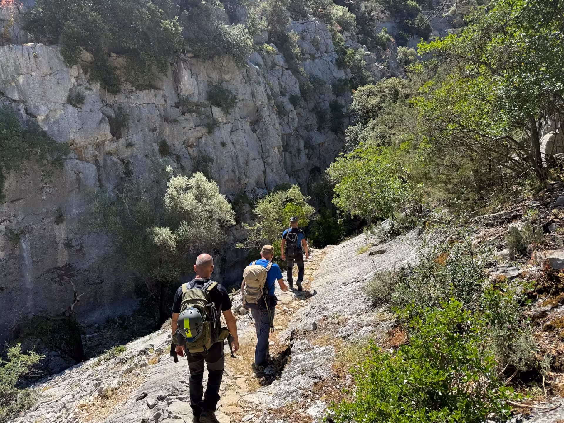 Hikers navigate a rocky trail surrounded by lush greenery and steep cliffs near Sa Nurre de Su Hoda Cave in Dorgali.
