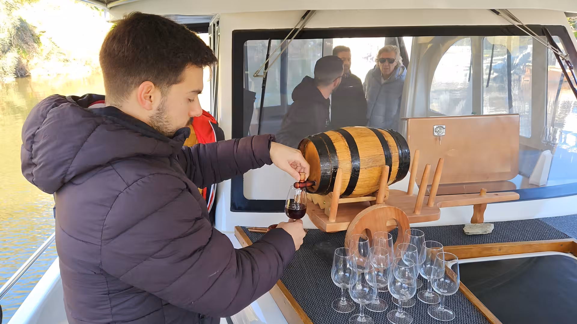 Onboard Douro River cruise wine tasting, pouring port-style wine from a wooden barrel with glasses on deck