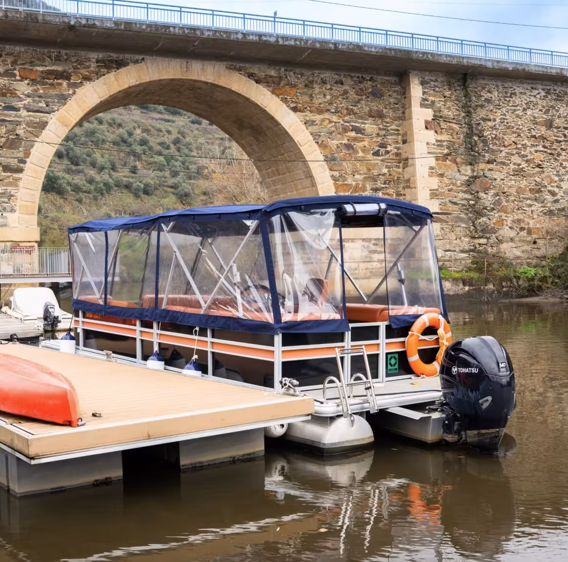 Private covered pontoon boat docked on the Douro River Valley near a historic stone arch bridge for a scenic cruise