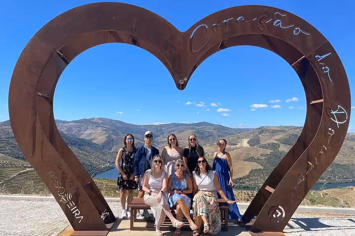 Happy tour group posing at the iconic Coracao do Douro heart sculpture with stunning Douro Valley vineyard views from Porto.