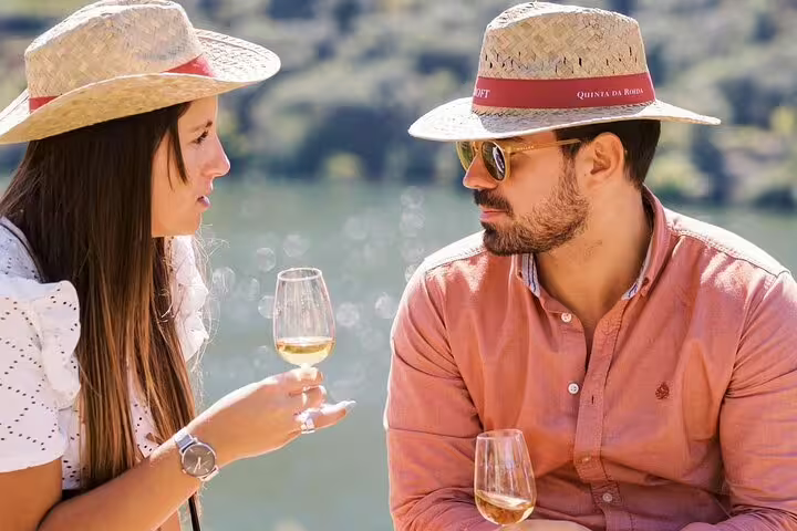 Couple enjoying wine tasting outdoors with Douro Valley views, wearing straw hats.