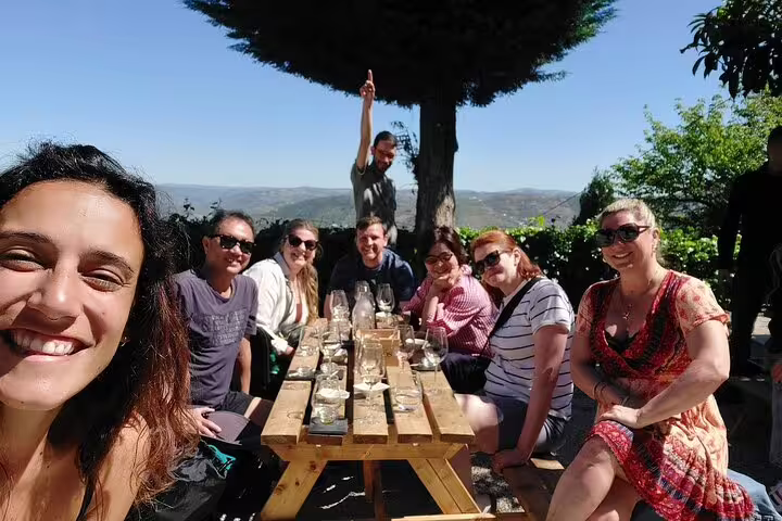 Group of happy tourists enjoying a Douro wine tasting tour at an outdoor picnic table with stunning vineyard views.