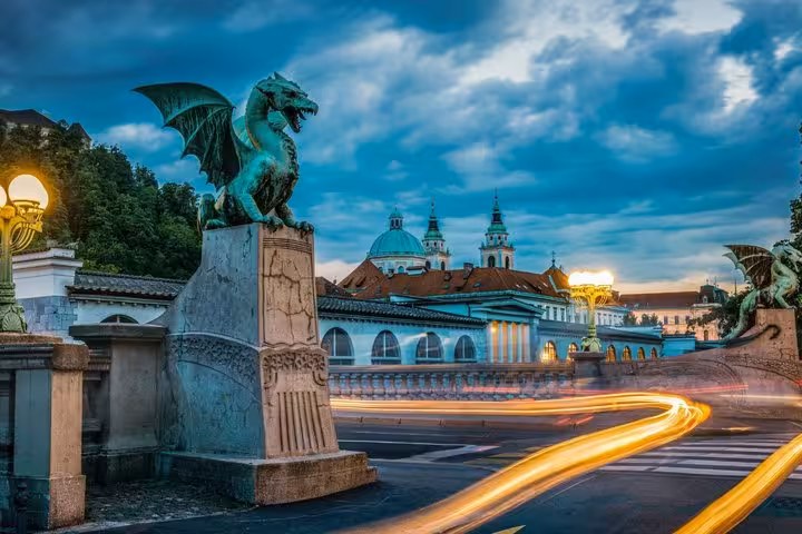 Dragon Bridge bronze dragon statue at dusk with light trails and Ljubljana Cathedral in background