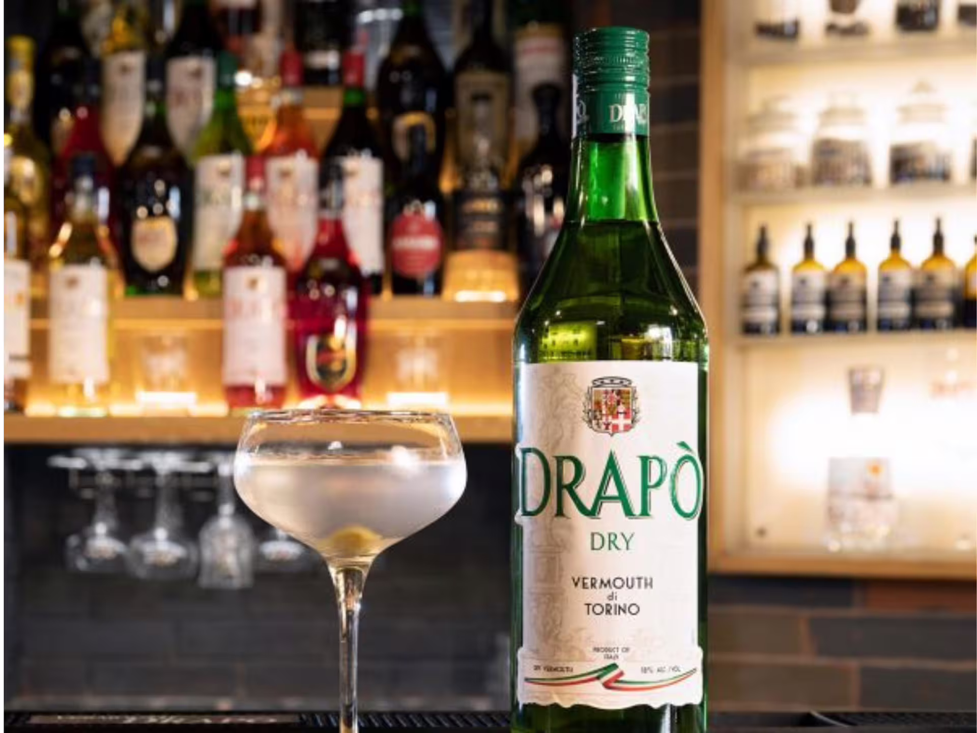 Explore Drapò Dry Vermouth in Turin, featuring a bottle and cocktail glass set against a vibrant bar backdrop.
