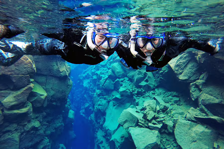 Snorkelers explore the crystal-clear waters of Silfra, Iceland, in drysuits, surrounded by stunning underwater rock formations.