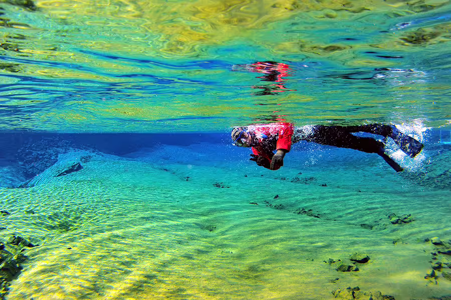 Snorkeler in a drysuit explores the crystal-clear waters of Silfra fissure, capturing underwater photos in Iceland's vivid landscape.