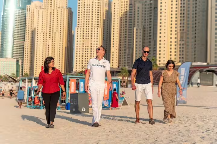 Group of tourists walk along Dubai Marina beach on a guided city tour, skyscrapers and promenade visible in the golden sunset.