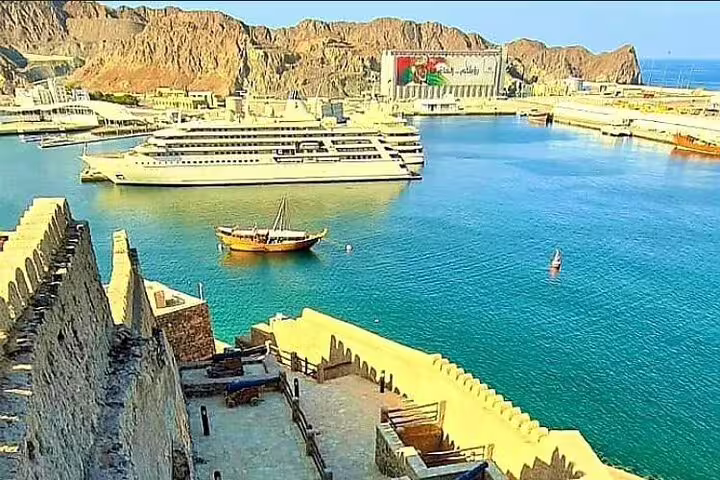 View of Muscat harbor with cruise ship and traditional boat, showcasing scenic Oman coastline for Dubai to Muscat transfer.