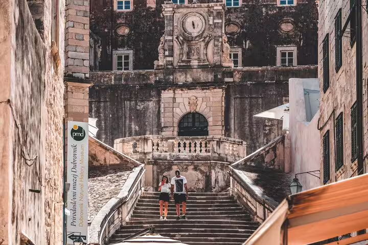 Two tourists descending historic stone stairs in Dubrovnik Old Town near Natural History Museum