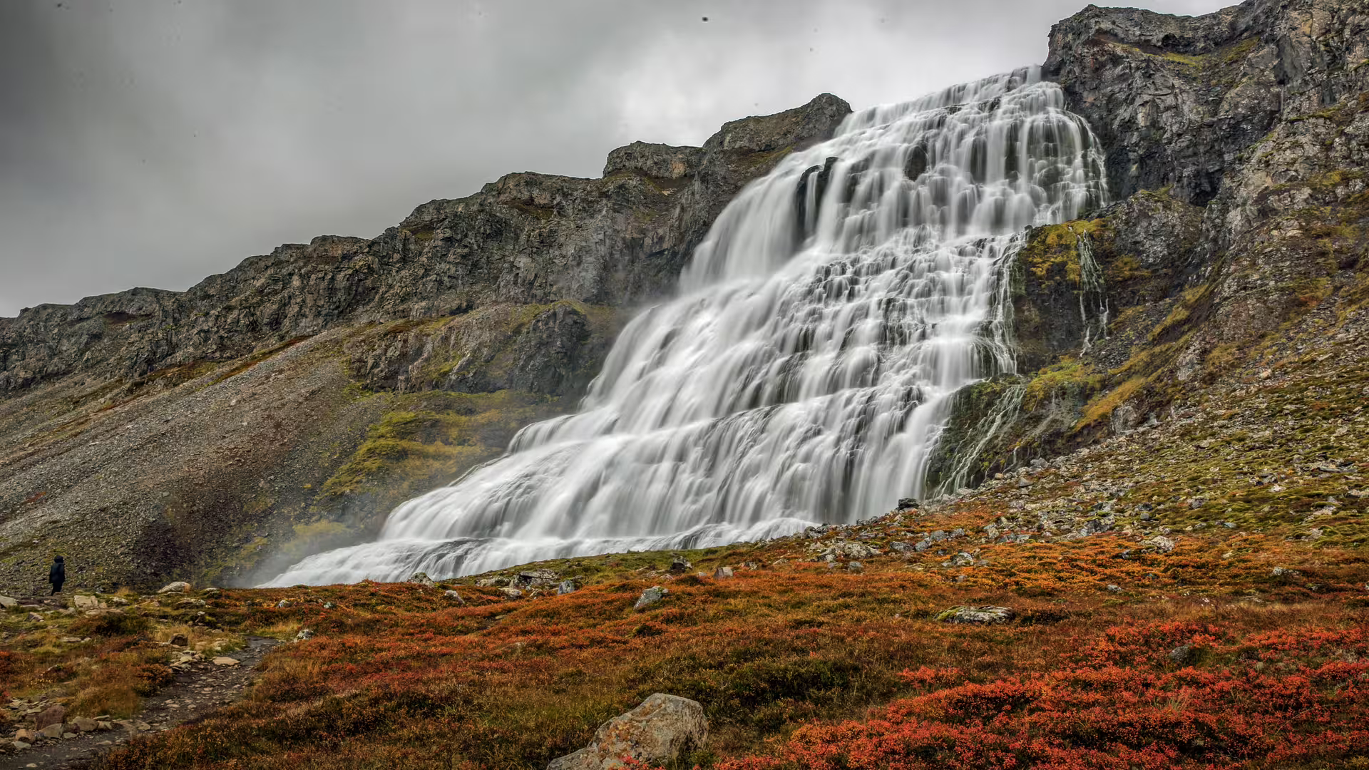 Dynjandi waterfall cascading down rocky cliffs with autumn red vegetation and a lone visitor in Isafjordur, Iceland
