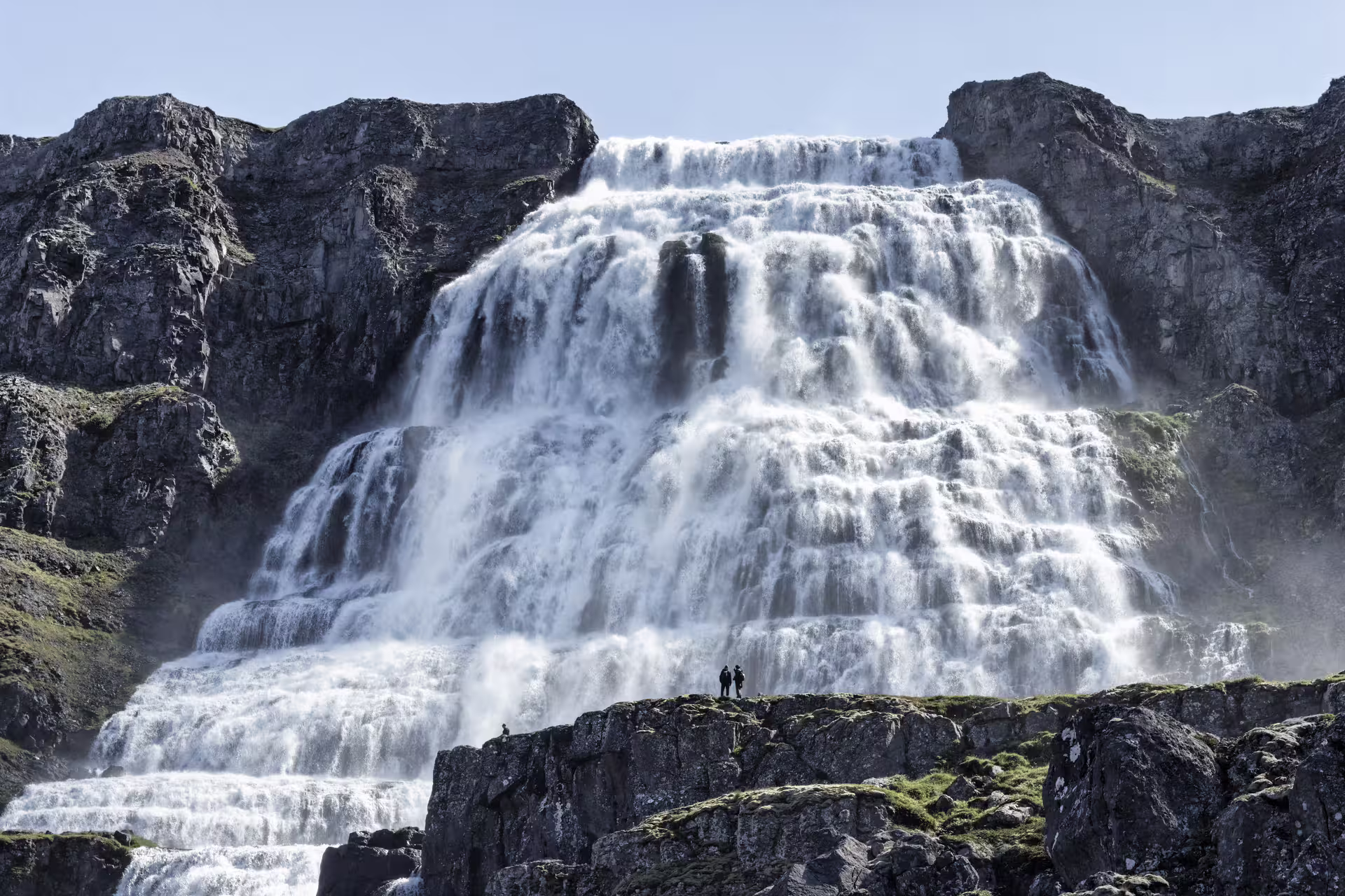 Two visitors standing on rocky ledge before the massive cascading tiers of Dynjandi Waterfall in Iceland