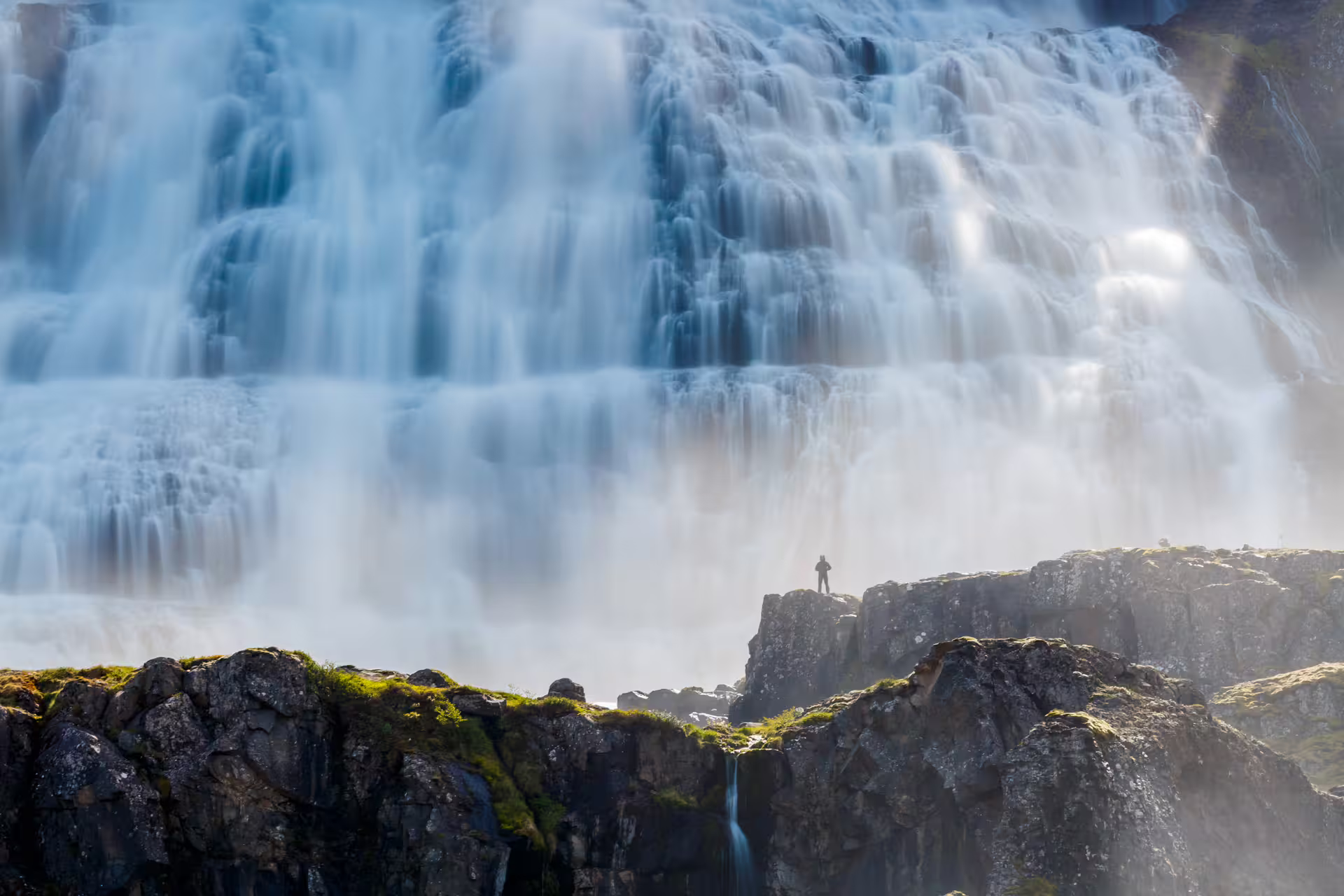 Tiny hiker silhouette standing on rocky cliff before the massive Dynjandi waterfall in Iceland