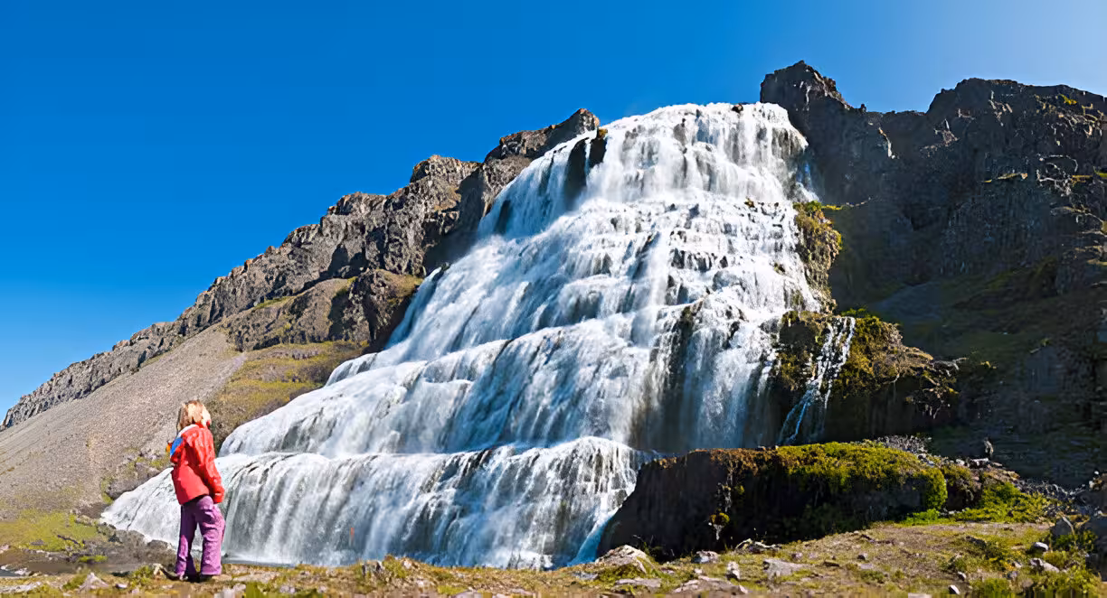 Hiker in red jacket standing before the cascading tiers of Dynjandi waterfall in Iceland's Westfjords