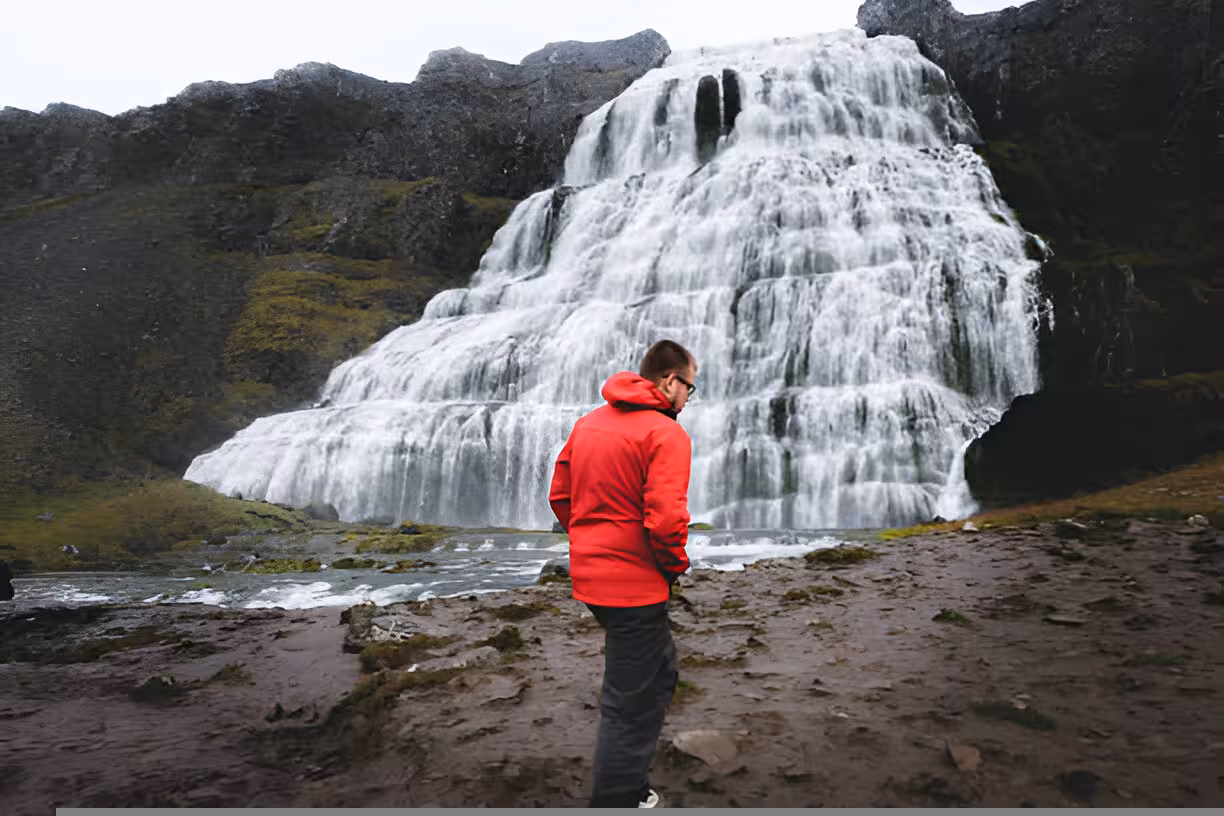 Visitor in red jacket standing before Dynjandi waterfall cascading down rocky cliffs in Iceland's Westfjords