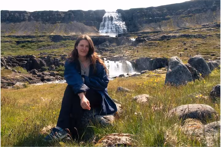 Woman sitting in grassy field before Dynjandi waterfall in Iceland's Westfjords on sunny day