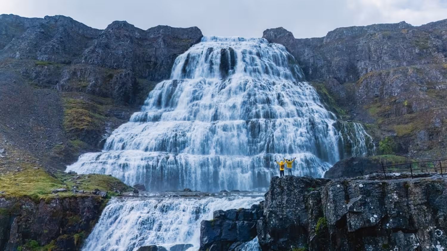 Two visitors in yellow jackets standing on rocks before the massive tiered Dynjandi waterfall in Iceland's Westfjords