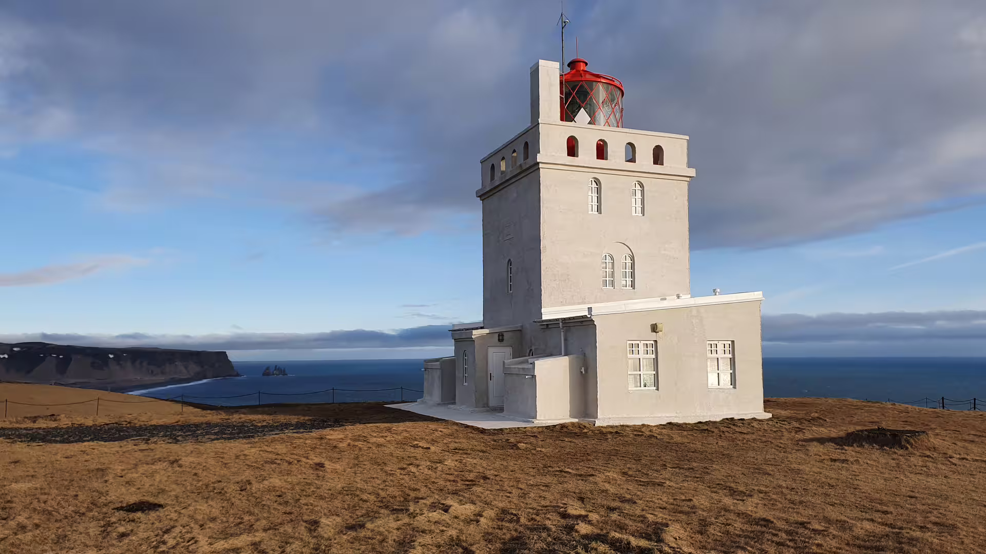 White lighthouse with red lantern on coastal cliffs overlooking black sand beach in South Iceland