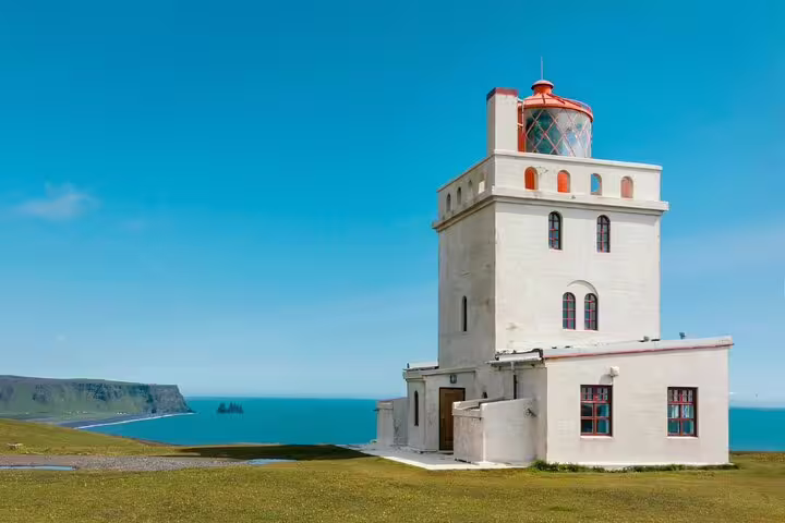 White lighthouse on Iceland's south coast cliffs overlooking turquoise ocean under clear blue sky