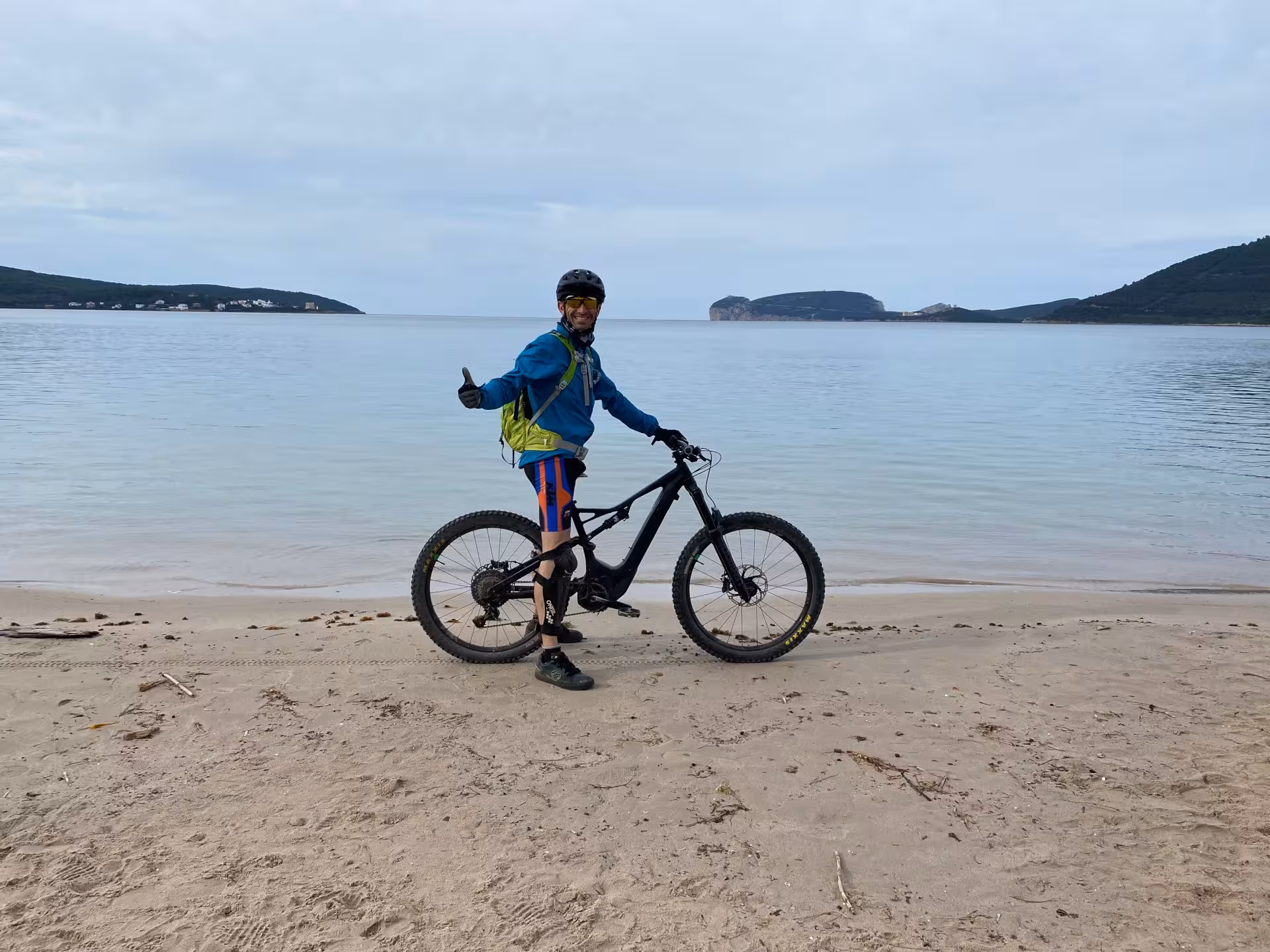 Cyclist on an e-bike posing on a sandy beach with a backdrop of the serene Alghero coastline and clear sea.