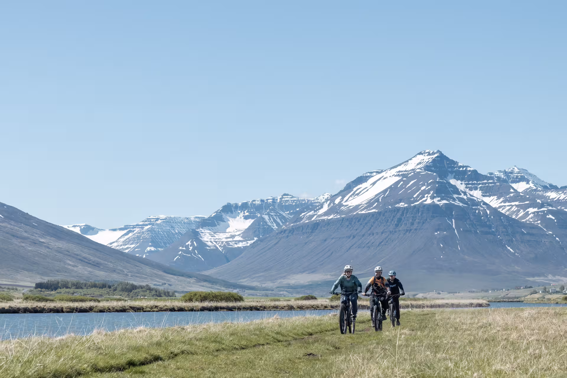 Three cyclists on e-bikes riding through green meadows with snow-capped mountains in Svarfaðardalur, Iceland