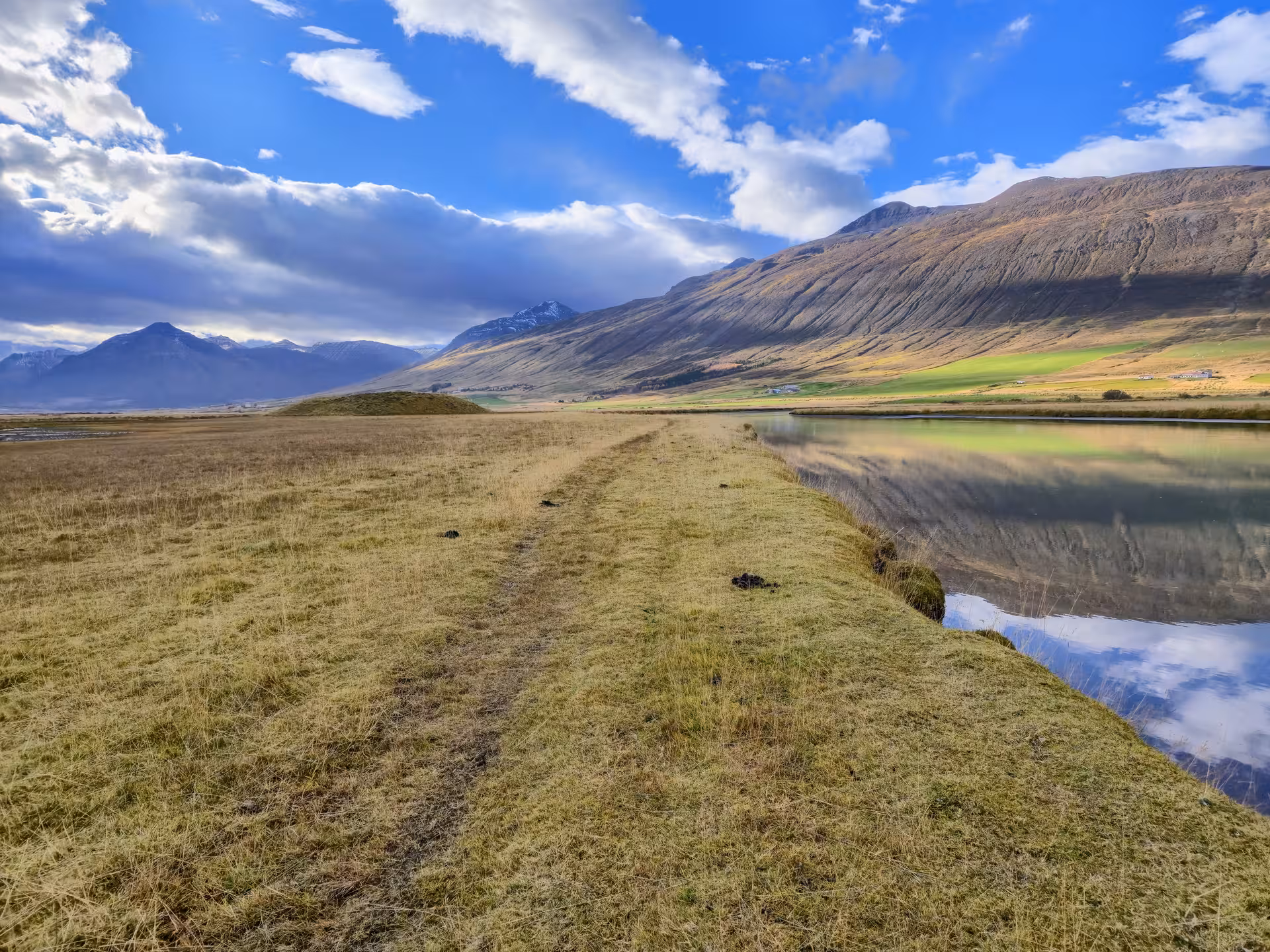 Grassy path alongside calm reflective canal with dramatic mountains and blue sky in Svarfaðardalur valley