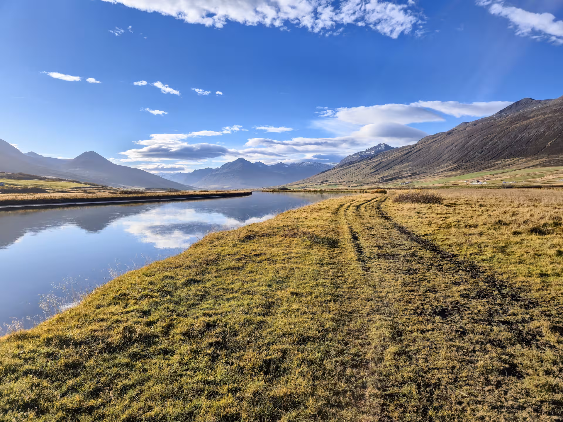 Scenic canal path through Svarfaðardalur valley with mountain reflections on calm water under blue sky