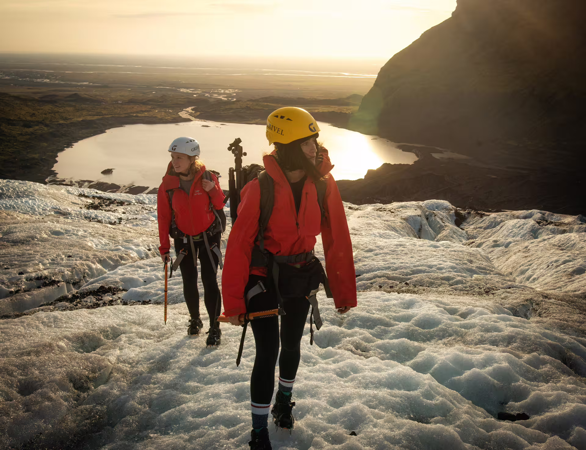 Two hikers in red jackets and helmets trek across an Iceland glacier at golden sunrise on an early bird ice cave tour