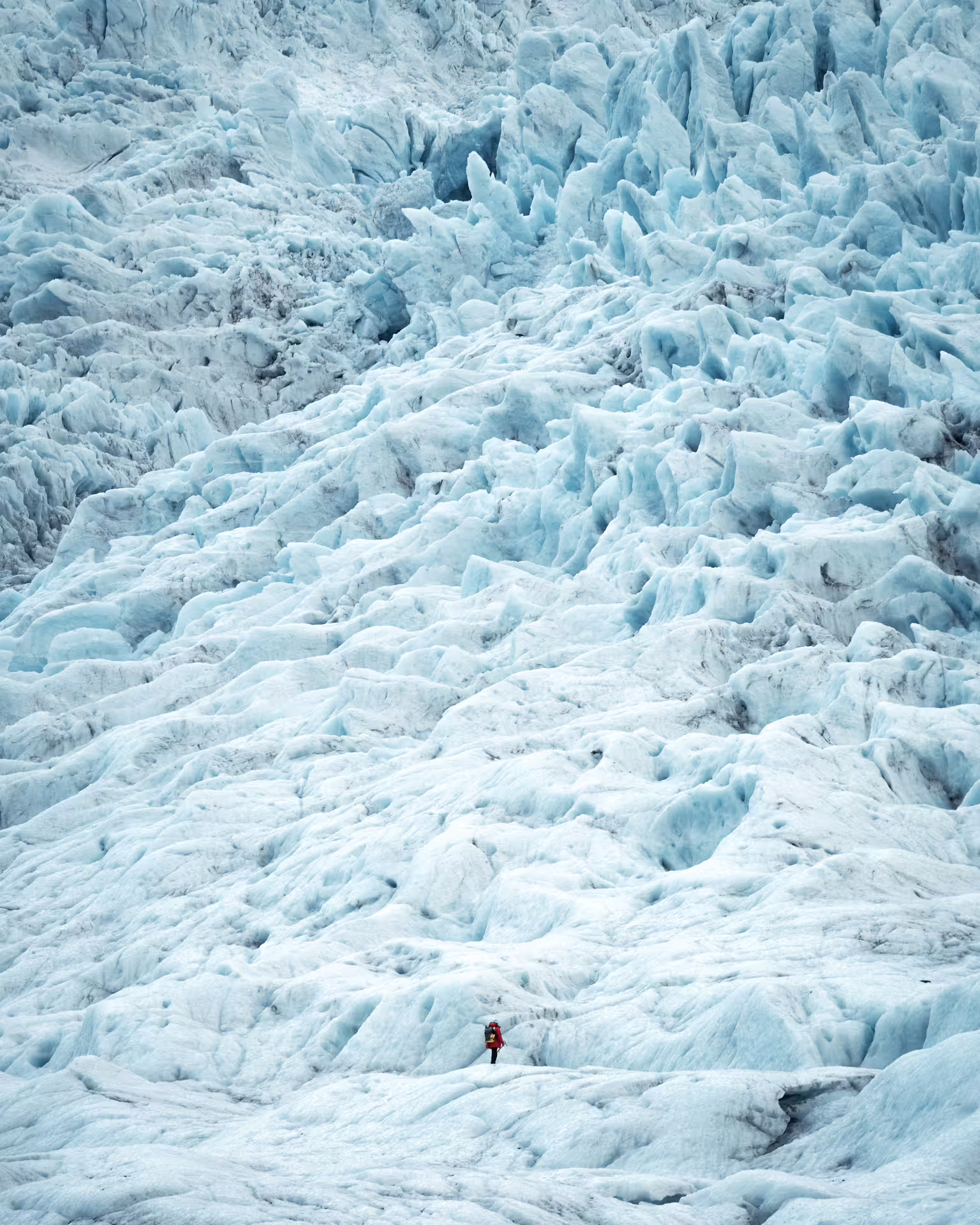 A lone hiker in red stands dwarfed by a vast blue glacial landscape on an early bird Iceland ice cave tour