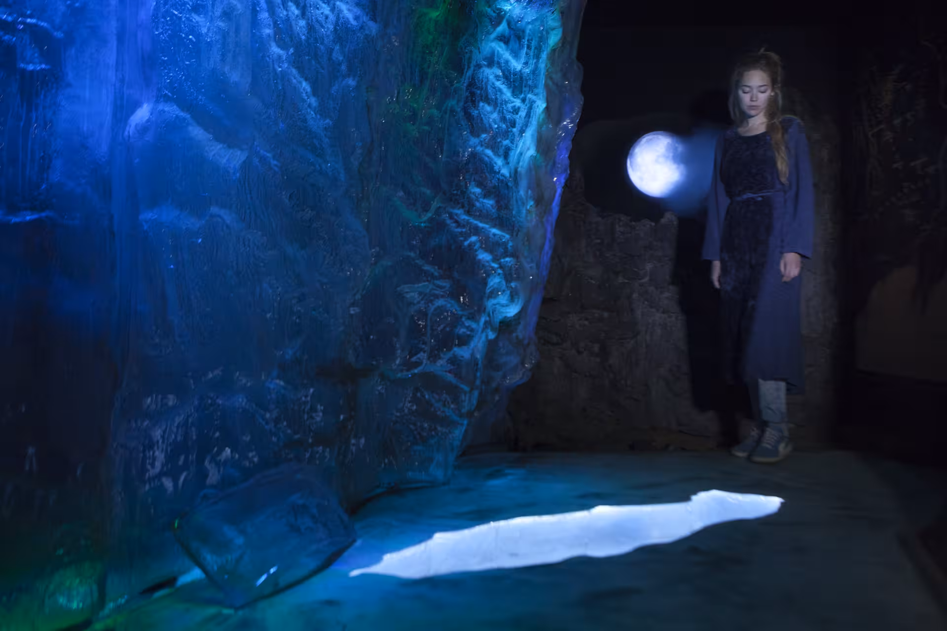 Woman in dark cave with glowing orb light beside blue illuminated ice wall in East Iceland