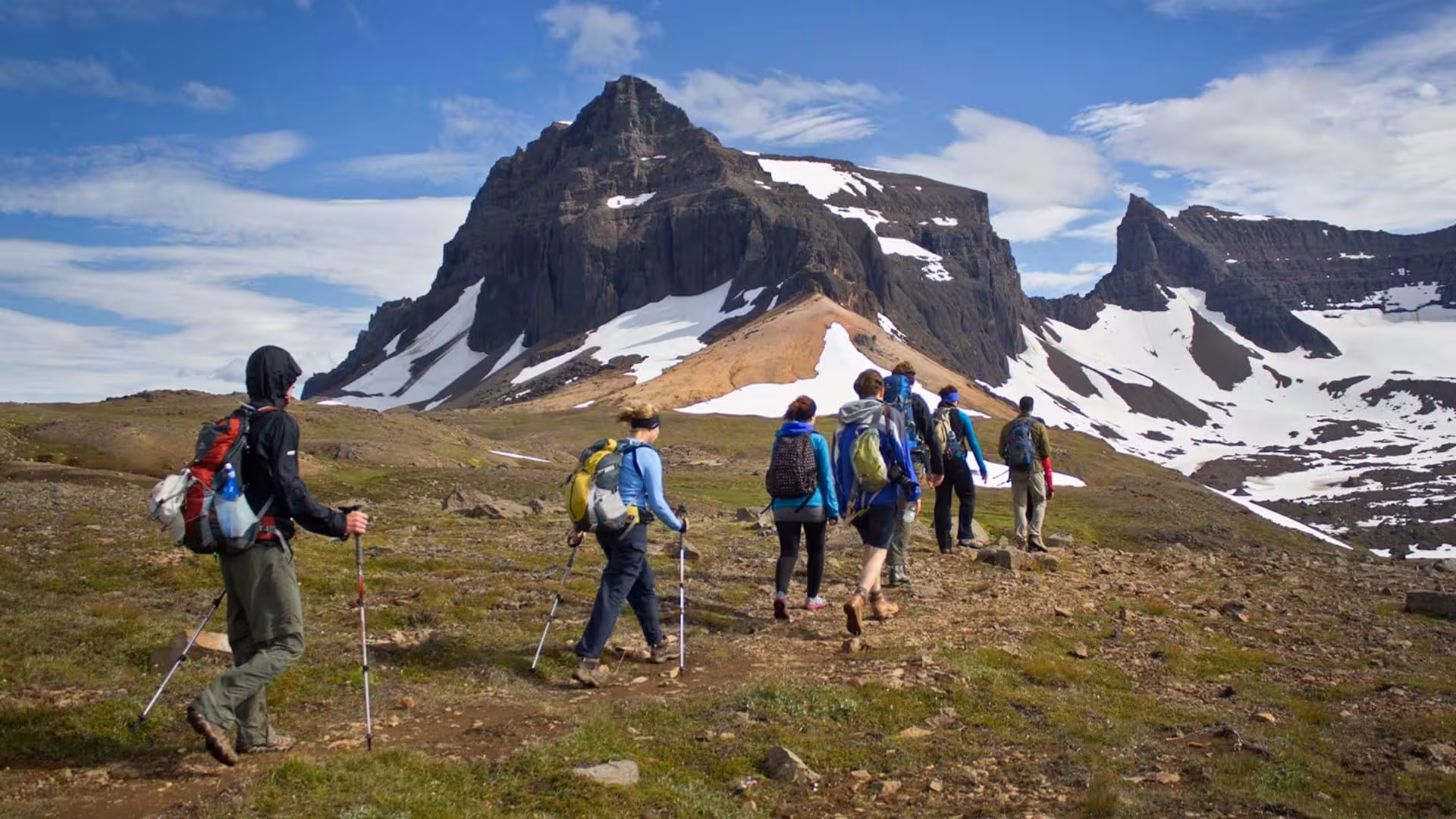 Group of hikers with backpacks and trekking poles ascending toward snow-capped mountains on East Iceland remote fjords tour