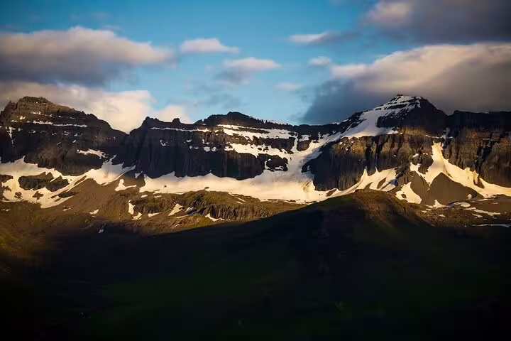 Snow-capped rugged mountain range with green valley in East Iceland under partly cloudy sky