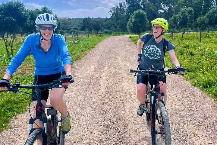 Two cyclists smile while riding e-bikes on a sunny backroad trail surrounded by greenery in Cascais and Sintra.