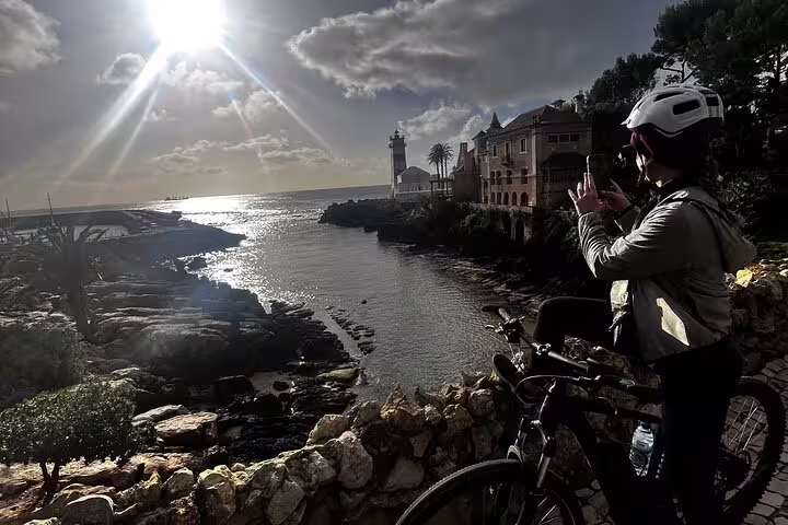 Cyclist pauses to capture stunning ocean views at Cascais, with the sun setting over the coastline and lighthouse.