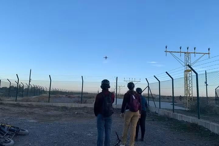 E-fat bike riders on Malaga off-road tour near airport fence, watching a plane over gravel track at sunset