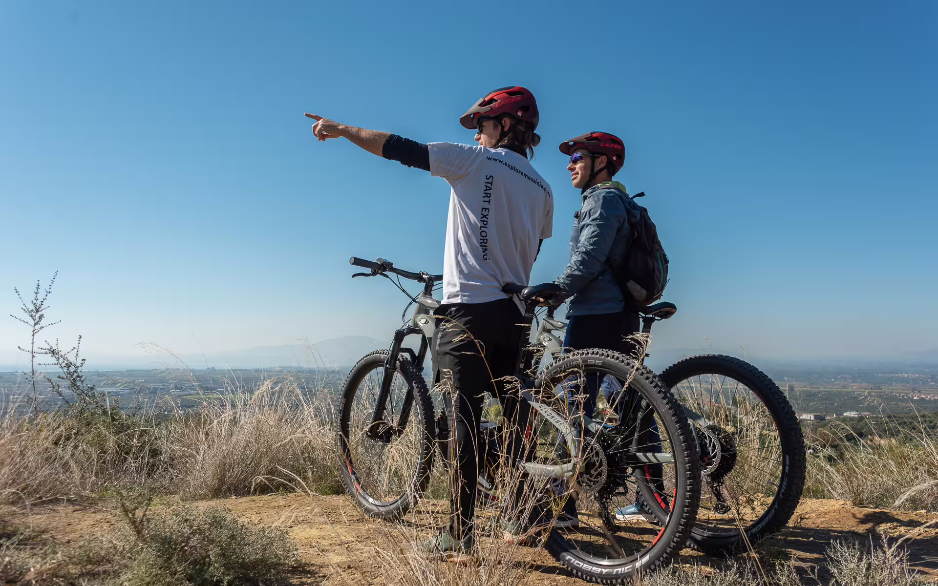 Guide pointing out scenery during an E-Bike Kalamata Olive Routes tour, with mountain bikes and Messinia views