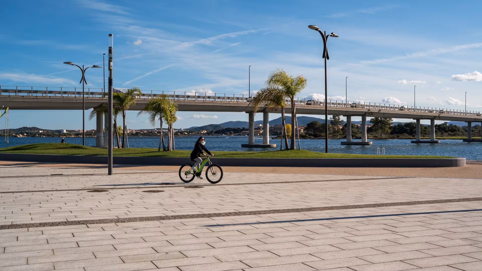 Cyclist riding an e-bike near a picturesque bridge and waterfront in Olbia, Sardinia, offering a relaxing holiday experience.