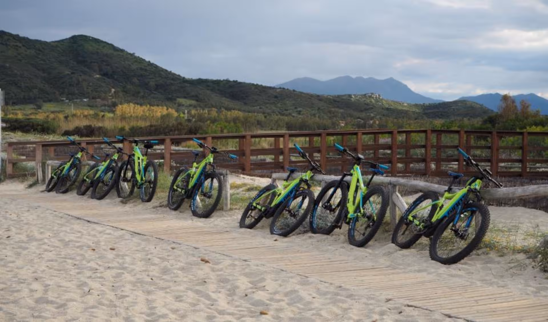 Line of e-bikes on a sandy path in Olbia, Sardinia, with lush green hills in the background.