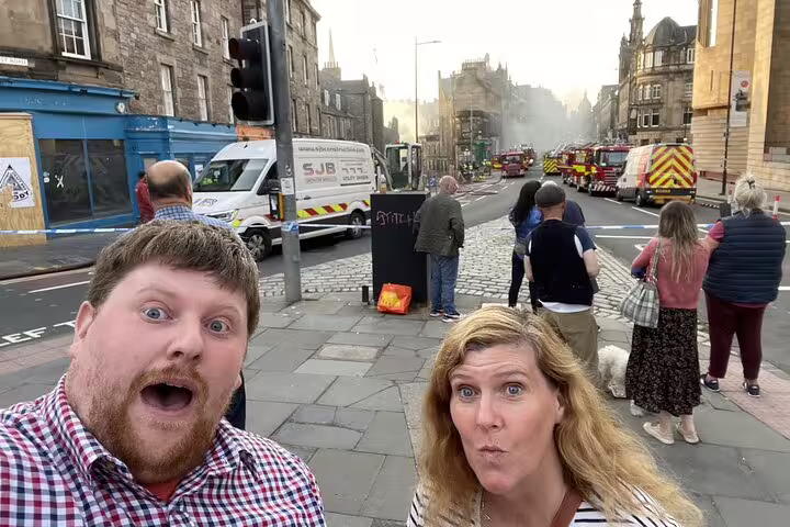 Excited couple taking selfie on Edinburgh cobblestone street with fire trucks and smoke in background
