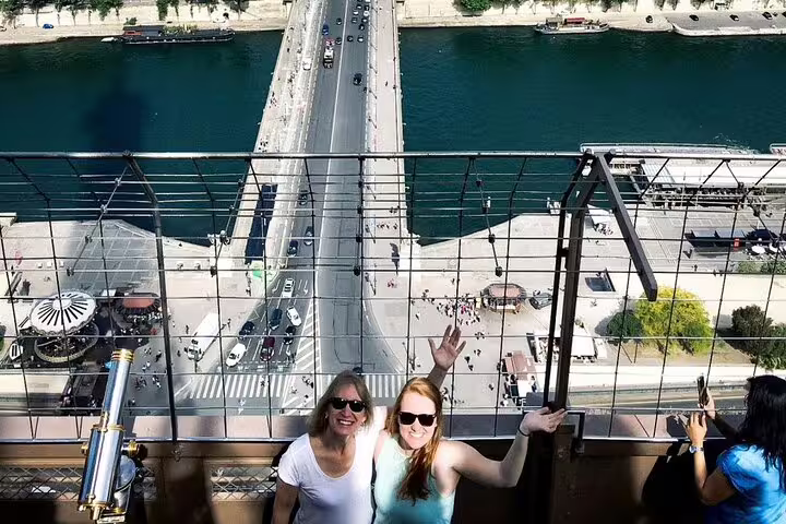 Two women smiling on Eiffel Tower observation deck with aerial view of Paris bridge and Seine River below