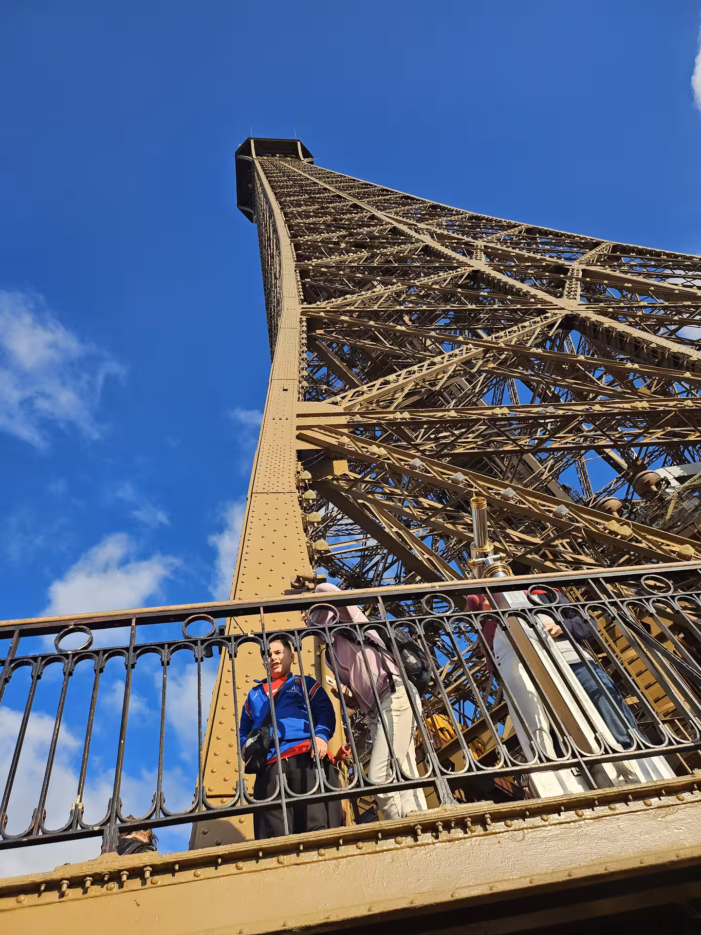 Tourists on Eiffel Tower first floor balcony with iron lattice structure soaring against blue sky, Paris