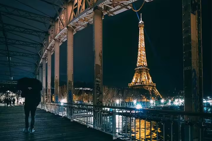 Eiffel Tower illuminated at night viewed from Bir-Hakeim Bridge with a silhouetted figure holding umbrella