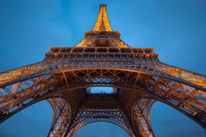 Eiffel Tower illuminated at dusk, dramatic upward view of iron lattice structure glowing golden against blue sky
