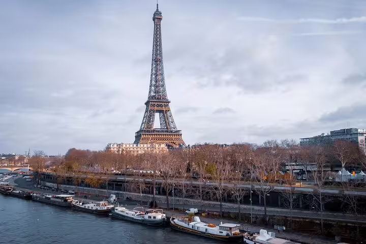 Eiffel Tower rising above the Seine River with boats docked along the Paris riverbank on a cloudy day