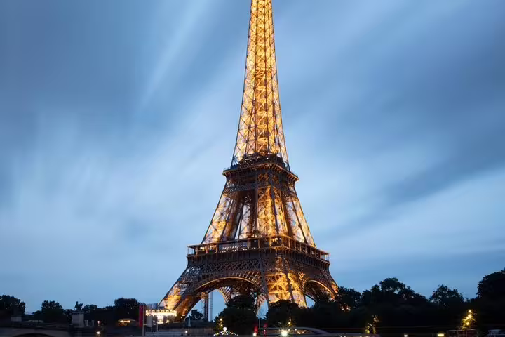 Eiffel Tower illuminated at dusk with glowing golden lights against a blue cloudy sky in Paris
